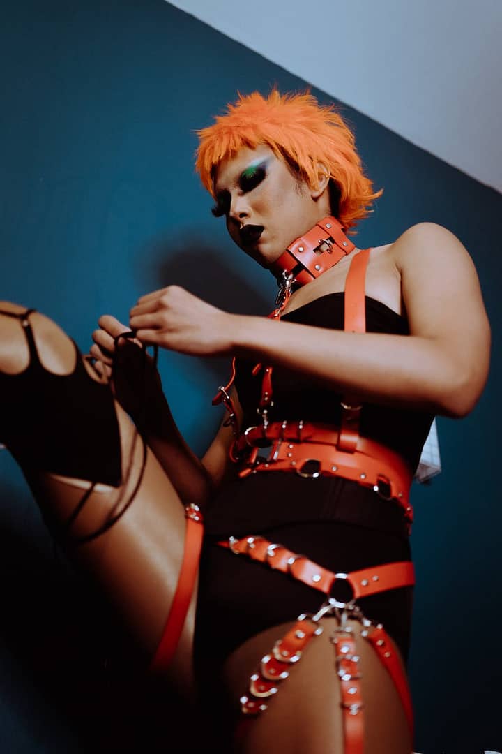 seductive ethnic transgender man in bdsm outfit tying shoelaces in room